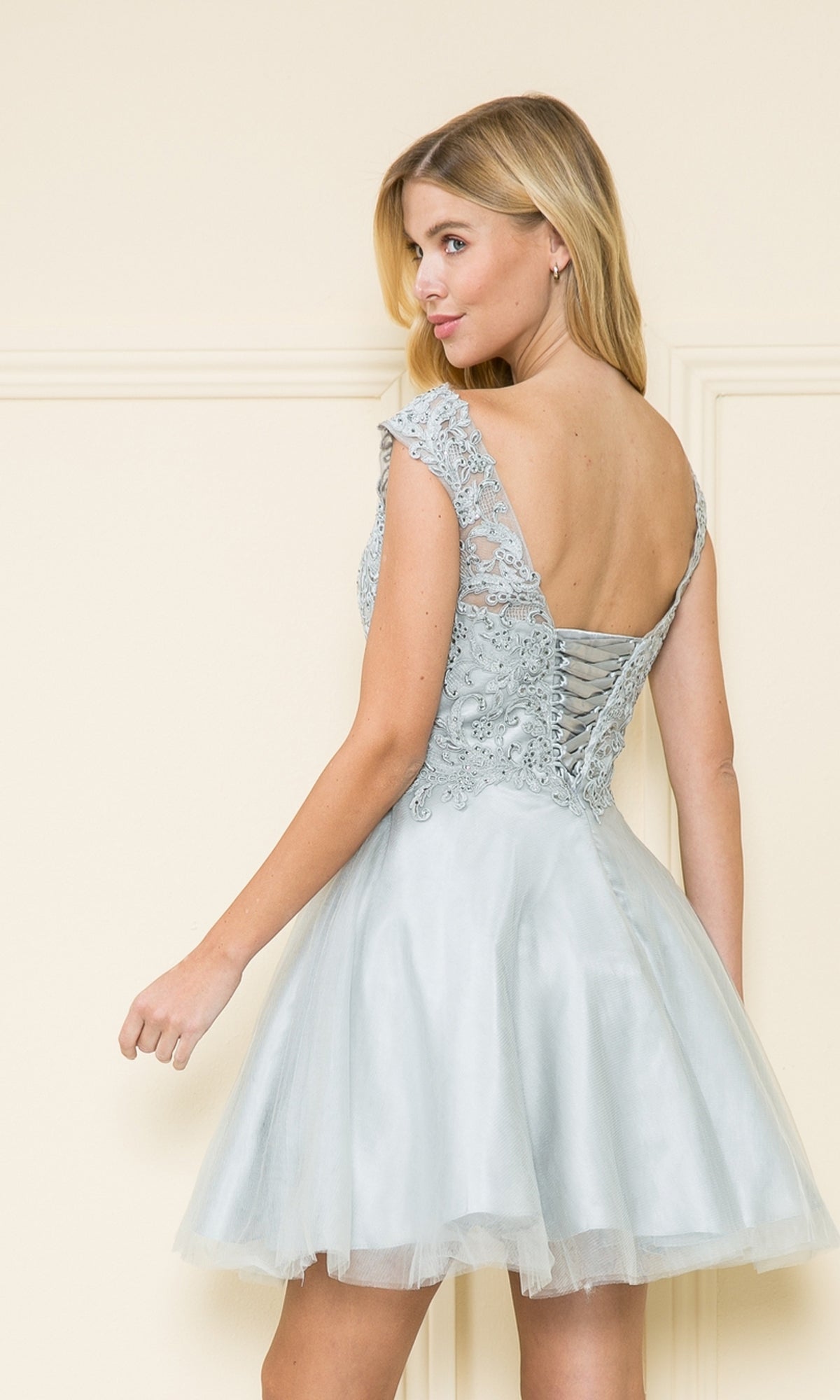 A woman in a Lace-Bodice Short Babydoll Homecoming Dress 8902 stands with her back turned, highlighting the lace-up detail and flattering a-line silhouette of this charming party dress.
